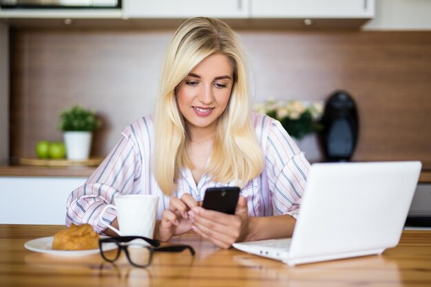 Zdjęcie kobieta w piżamie pijąca kawę i używająca smartfona i laptopa w kuchni