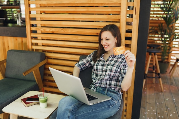 Kobieta w plenerze ulica letnia kawiarnia drewniana kawiarnia siedząca pracująca na komputerze przenośnym, trzymaj kartę kredytową banku relaksując się w czasie wolnym
