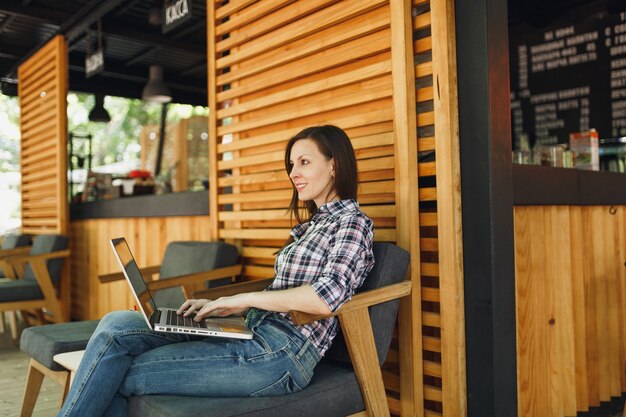 Kobieta w plenerze ulica letnia kawiarnia drewniana kawiarnia siedząca w codziennych ubraniach, pracująca na nowoczesnym komputerze typu laptop pc pc