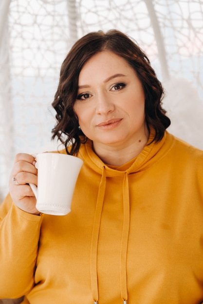 Kobieta W Pomarańczowej Bluzie Siedzi Na Niezwykłym Krześle I Pije Kawę W Domu.