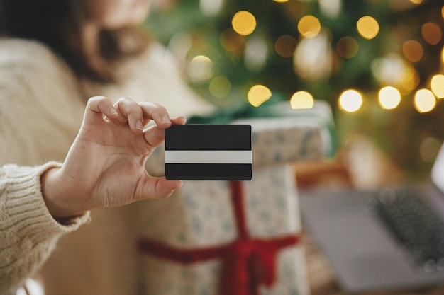 Kobieta w przytulnym swetrze, trzymająca kartę kredytową z bliska i stylowe prezenty świąteczne w świątecznie udekorowanym pokoju bożonarodzeniowym ze światłami Świąteczne zakupy online i wyprzedaże w Czarny Piątek
