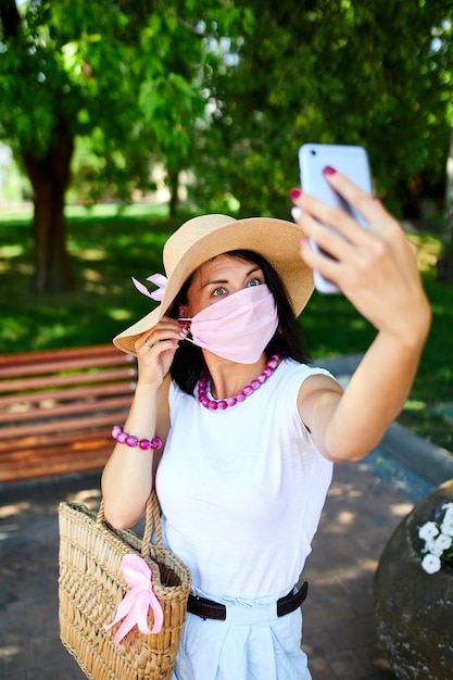 Kobieta W Różowej Masce Medycznej W Parku Robi Selfie Przez Telefon Komórkowy, Kobieta Z Ochroną Dróg Oddechowych Jest Na Zewnątrz Podczas Rozmowy I Czatu Wideo Przez Kamerę Internetową Na Smartfonie