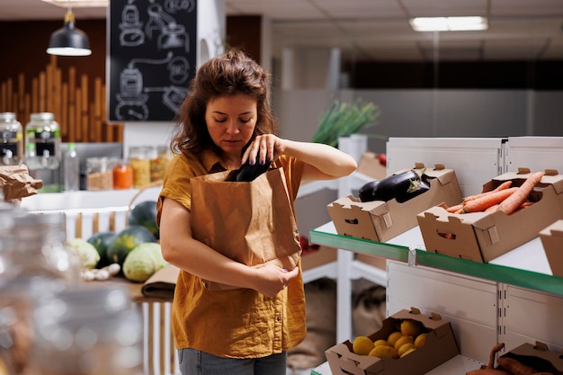 Zdjęcie kobieta w sklepie zero waste kupująca naturalne warzywa uprawiane na farmie i zbierająca dojrzałe bakłażany. klient w lokalnym sklepie spożywczym wolnym od plastiku, który chce kupić zdrową żywność przy użyciu biodegradowalnej torby papierowej