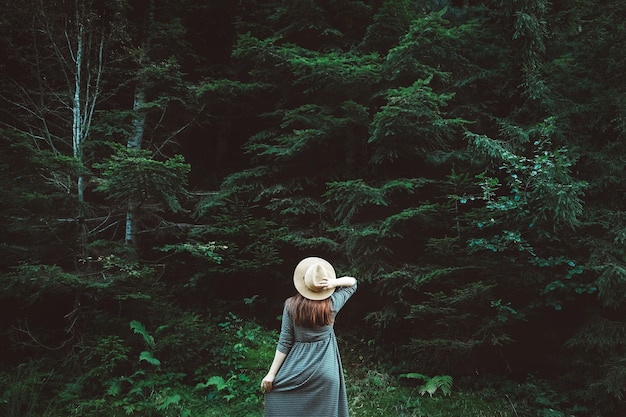 Kobieta w słomkowym kapeluszu i sukience stoi na tle zielonego lasu i drzew