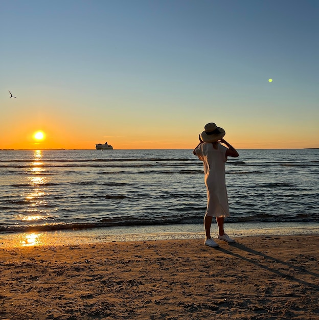 kobieta w słomkowym kapeluszu spacerująca po plaży na morzu o zachodzie słońca