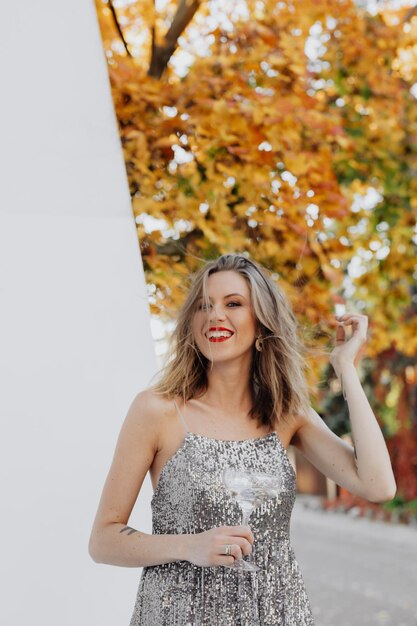 Zdjęcie kobieta w srebrnej sukience pozuje przed białą ścianą z jesiennymi liśćmi.