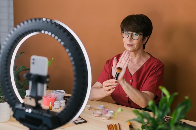 Kobieta w średnim wieku, blogerka lub influencerka zajmująca się urodą, nagrywająca wideo z produktami do makijażu w pokoju w domu u