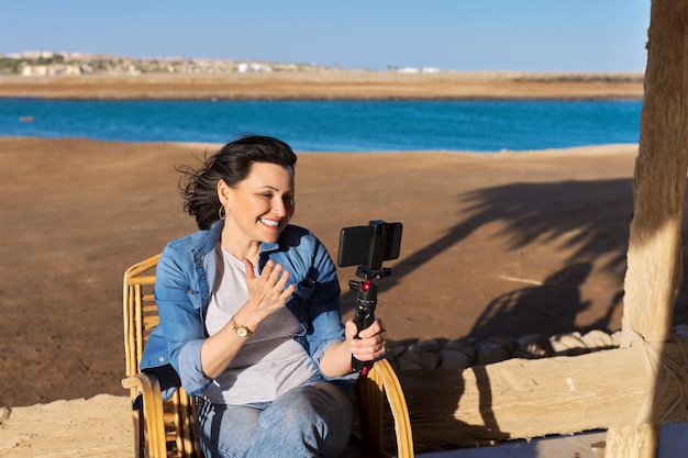 Kobieta W średnim Wieku, Patrząca Na Kamerę Internetową Smartfona, Rozmawiająca, Nagrywająca Wideo Na Piaszczystej Plaży