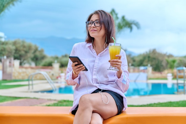 Kobieta w średnim wieku relaksuje się przy basenie za pomocą słuchawek i smartfona