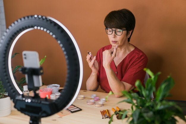 Kobieta w średnim wieku rozmawia o kosmetykach z cieniami do makijażu i paletą różu i pędzlami podczas nagrywania filmu Dojrzała kobieta robi film na swoim blogu o kosmetykach