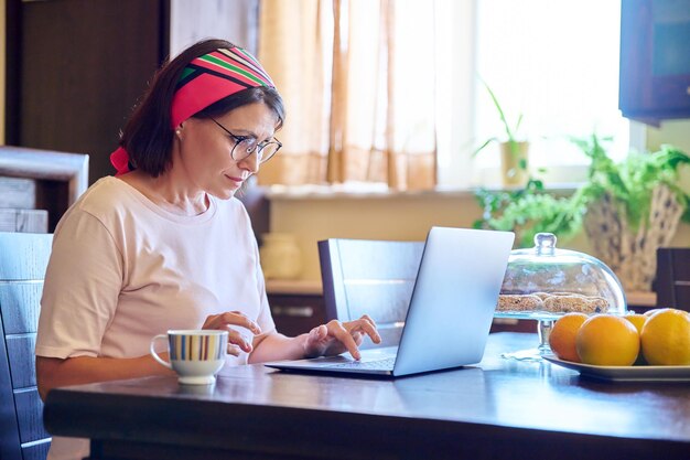 Kobieta w średnim wieku siedzi w domu w kuchni z laptopem i filiżanką kawy