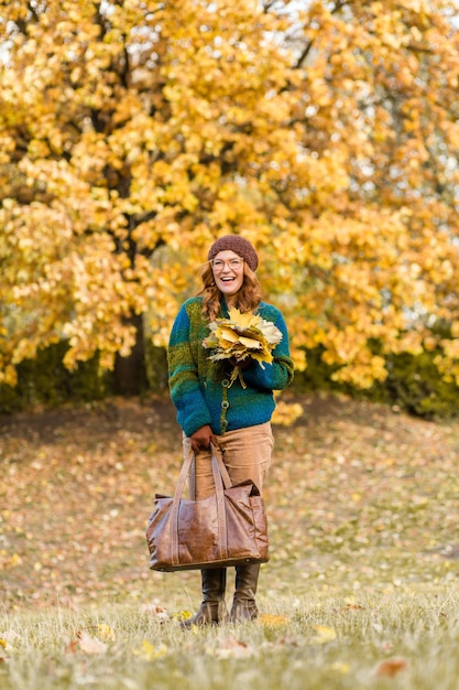 Kobieta w średnim wieku spacerująca po parku i uśmiechająca się z liśćmi klonu Szczęśliwa dama w brązowym kapeluszu i kurtce trzymająca skórzaną kurtkę
