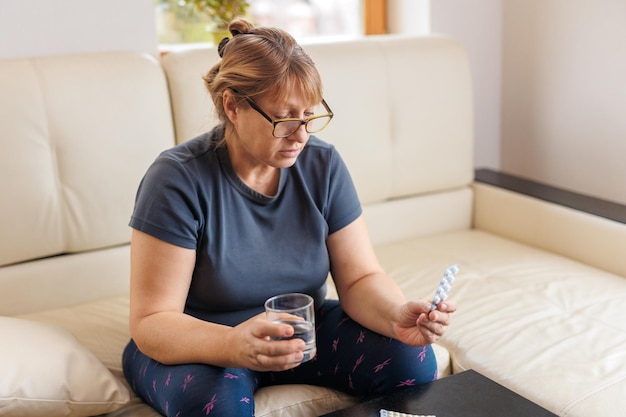 Kobieta w średnim wieku trzyma blister z tabletkami i czyta instrukcje medyczne, siedząc na kanapie