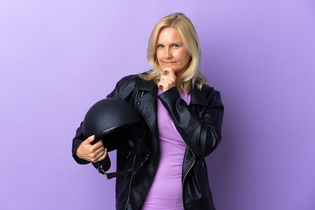 Kobieta w średnim wieku trzyma kask motocyklowy na białym tle na fioletowej ścianie, mając wątpliwości, patrząc w górę