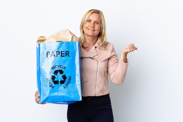 Kobieta w średnim wieku trzyma worek recyklingu pełen papieru do recyklingu na białym tle na białej ścianie, wskazując na bok, aby przedstawić produkt
