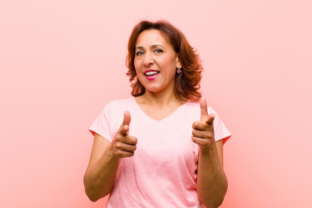 Kobieta w średnim wieku, uśmiechając się z pozytywnym, udanym, szczęśliwym nastawieniem, wskazując, co pistolet znak rękami na różowej ścianie