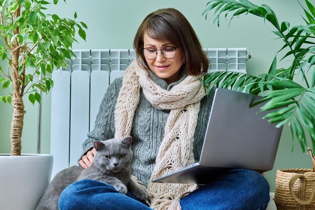 Kobieta w średnim wieku w ciepłych ubraniach z kotem za pomocą laptopa w domu