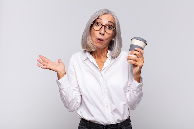 Kobieta w średnim wieku wyglądająca na zaskoczoną i zszokowaną, z opuszczoną szczęką, trzymając przedmiot z otwartą ręką na bocznej koncepcji kawy