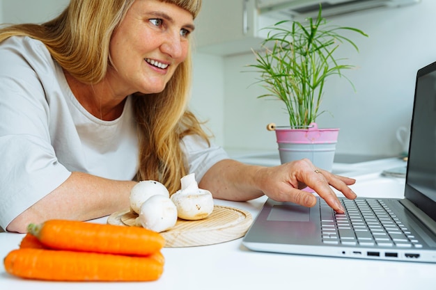 kobieta w średnim wieku z długimi blond włosami gotuje w kuchni i z zainteresowaniem patrzy na ekran laptopa