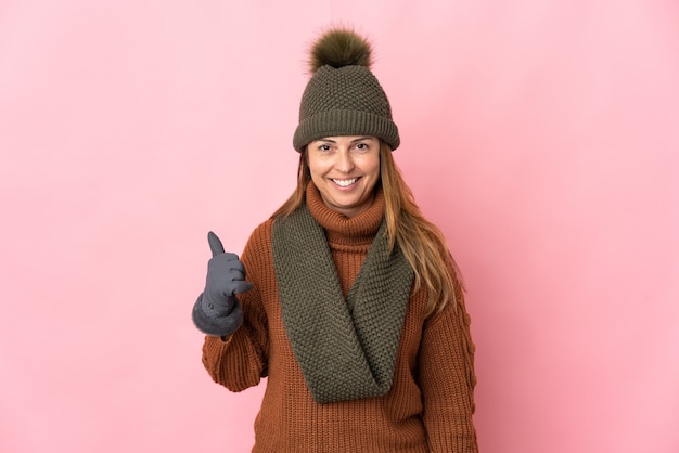 Kobieta w średnim wieku z kapeluszem zimowym na białym tle na różowej ścianie, wskazując w bok, aby przedstawić produkt
