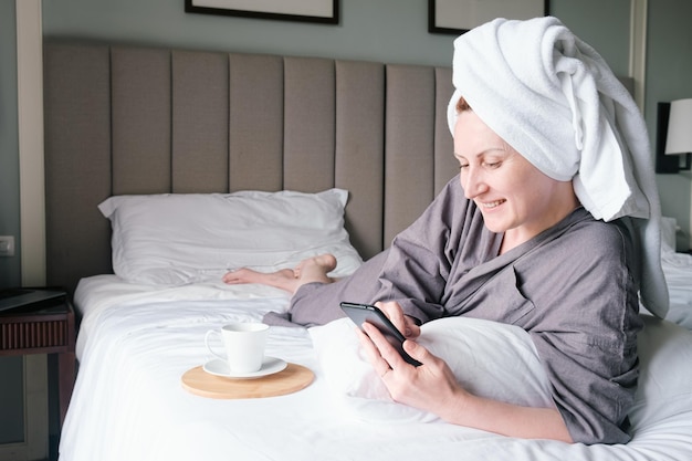 Kobieta w średnim wieku z ręcznikiem na głowie leży na łóżku uśmiechając się i patrząc na ekran smartfona