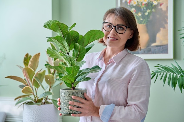 Kobieta w średnim wieku z rośliną ficus lyrata w doniczce uśmiechnąca się kobieta patrząca do kamery w domu Opieka nad roślinami domowymi koncepcja ludzi przyrody