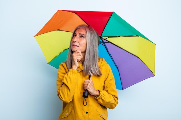 Kobieta w średnim wieku z siwymi włosami, uśmiechnięta ze szczęśliwym, pewnym siebie wyrazem twarzy z ręką na brodzie. koncepcja parasola i deszczu