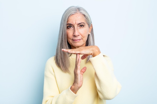 Kobieta w średnim wieku z siwymi włosami wyglądająca poważnie, surowo, zła i niezadowolona, robiąc znak czasu na znak
