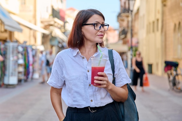 Kobieta w średnim wieku ze szklanką świeżego soku w dłoniach spacerująca po mieście