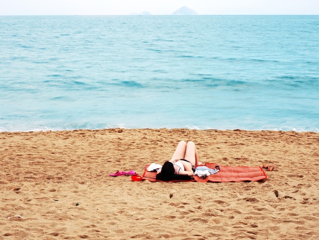 Zdjęcie kobieta w stroju kąpielowym bikini połóż się na czerwonej macie do opalania i zrelaksuj się na piaszczysto-niebieskiej plaży morskiej