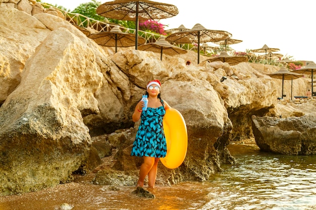 Kobieta w stroju kąpielowym i czapce Mikołaja na kamienistym brzegu morza z kółkiem do pływania i koktajlem podczas świątecznych wakacji