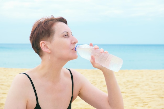 Kobieta w stroju kąpielowym siedzi na plaży i pije wodę z plastikowej butelki