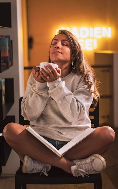 Kobieta w swetrze, patrząca w górę, popijając kawę trzymając książkę w księgarni