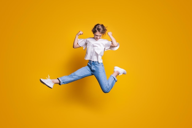 Kobieta w swobodnych dżinsach skacze z uniesionymi rękami i nogami na żółtym tle zwycięzca