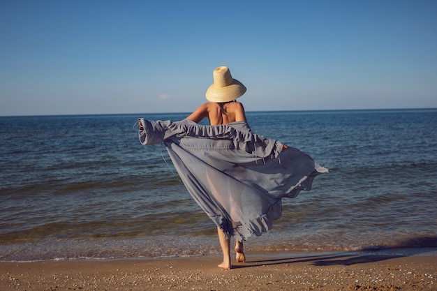 Kobieta w tunice, czarnym kostiumie kąpielowym i słomkowym kapeluszu stoi latem na plaży nad morzem na wakacjach