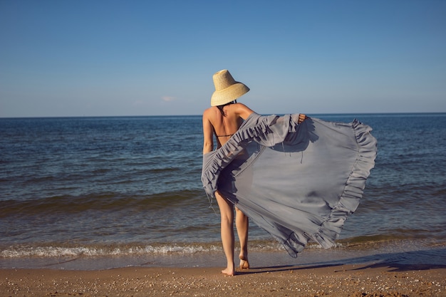 Kobieta w tunice, czarnym kostiumie kąpielowym i słomkowym kapeluszu stoi latem na plaży nad morzem na wakacjach