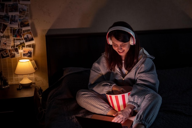 Kobieta w zestawie słuchawkowym siedzi na łóżku jedząc kukurydzę i oglądając film
