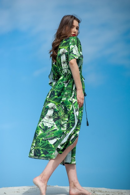 Kobieta w zielonej sukience z nadrukiem zielonych liści palmowych na dole i białej sukni z błękitnym niebem za nią.