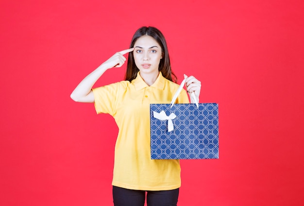 kobieta w żółtej koszuli trzyma niebieską torbę na zakupy i wygląda na zdezorientowaną i zamyśloną.