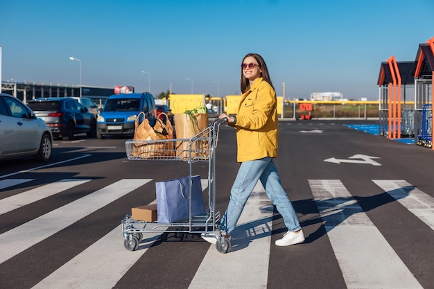 Kobieta w żółtej kurtce przechodzi przez przejście dla pieszych z centrum handlowego z wózkiem na zakupy z artykułami spożywczymi