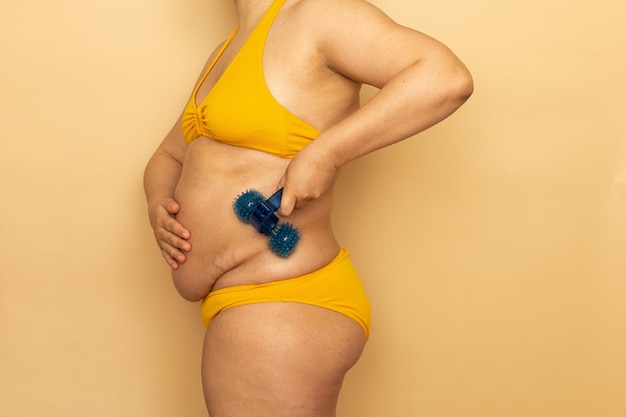Kobieta w żółtym stroju kąpielowym masuje gruby brzuch z niebieskim masażerem ciała widok z boku beżowe tło