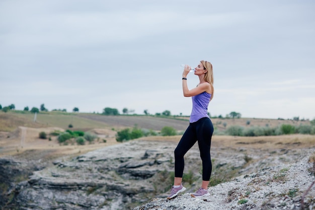 Kobieta Woda Pitna Po Sportach. na tle nieba. Sportowy styl życia, Atrakcyjna wysportowana dama spragniona wody pitnej po ciężkim treningu