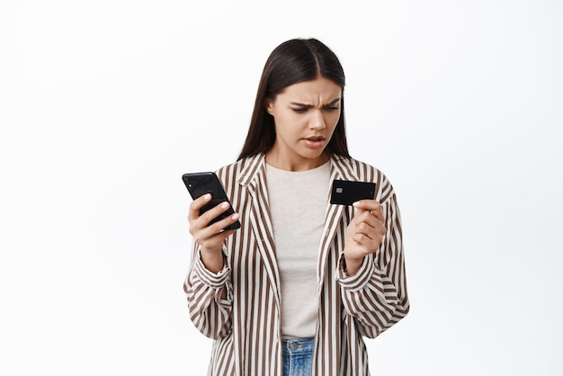 Kobieta wygląda na zdezorientowaną plastikową kartą kredytową podczas płacenia za zakupy online w aplikacji na smartfona lub przelewania pieniędzy za pomocą aplikacji na białym tle