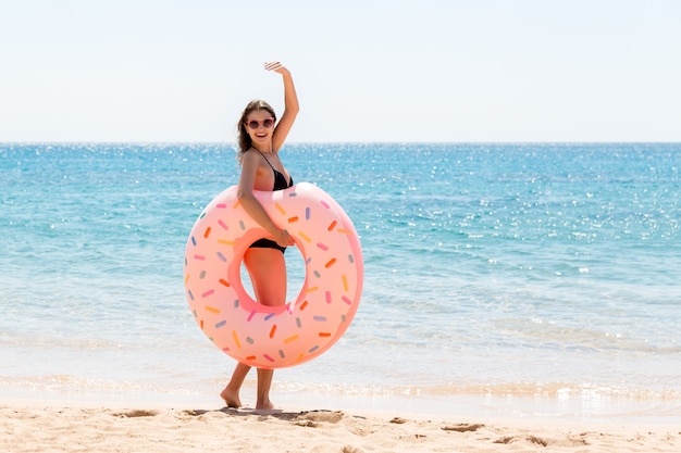 Kobieta wzywa do pływania w morzu i macha ręką. Dziewczyna relaksuje się z pączkiem na plaży i bawi się nadmuchiwanym pierścieniem. Letnie wakacje i koncepcja wakacji.