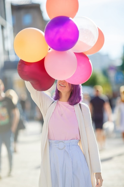 Kobieta z fiołkowymi włosami stoi w mieście i zakrywa jej twarz z balonami