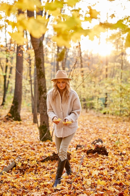 Kobieta z jesiennych liści w słonecznej przyrodzie, parku lub lesie. pani w kapeluszu chodzi