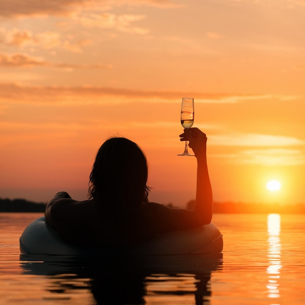 Zdjęcie kobieta z kieliszkiem szampana leżącego na gumowym pierścieniu w wodzie o zachodzie lub wschodzie słońca