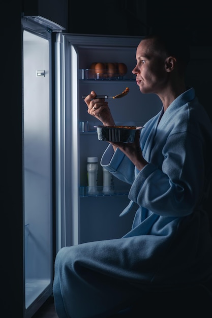 Kobieta z krótką fryzurą je w nocy w pobliżu lodówki Koncepcja nocnego apetytu Łysa szczupła kobieta zaspokaja głód