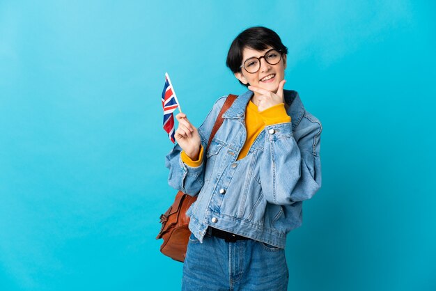 Kobieta z krótkimi włosami trzyma flagę Wielkiej Brytanii na białym tle na niebieskiej ścianie szczęśliwa i uśmiechnięta