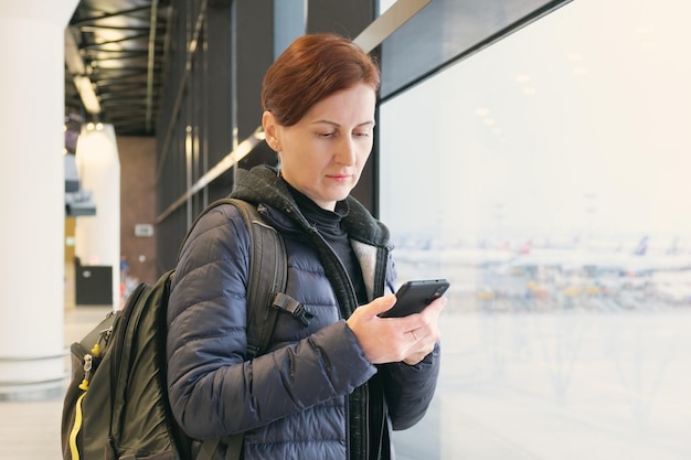 Kobieta z krótkimi włosami trzyma w dłoniach smartfon i patrzy na ekran, czekając na swój lot